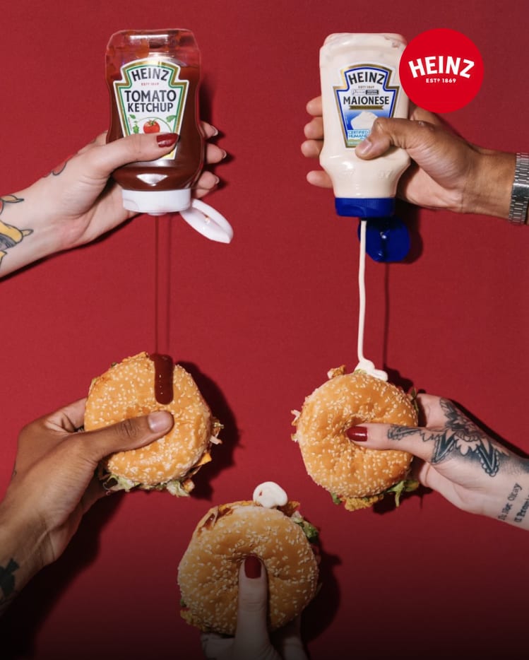 Hands squeezing Heinz ketchup and mayo onto sesame seed burgers against a red wall.