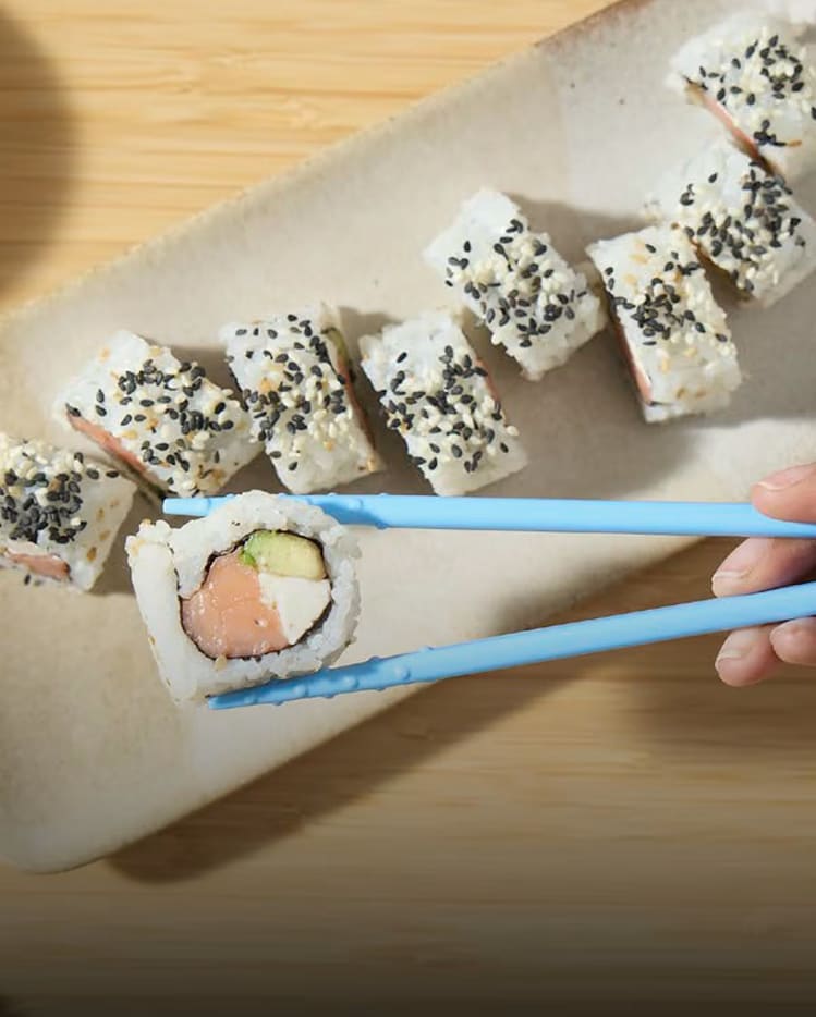 A close-up of a California roll with salmon, avocado, and Philadelphia cream cheese, held by chopsticks.