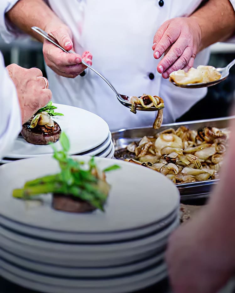 Chefs plating a dish with steak, asparagus and sauteed onions in a busy kitchen.
