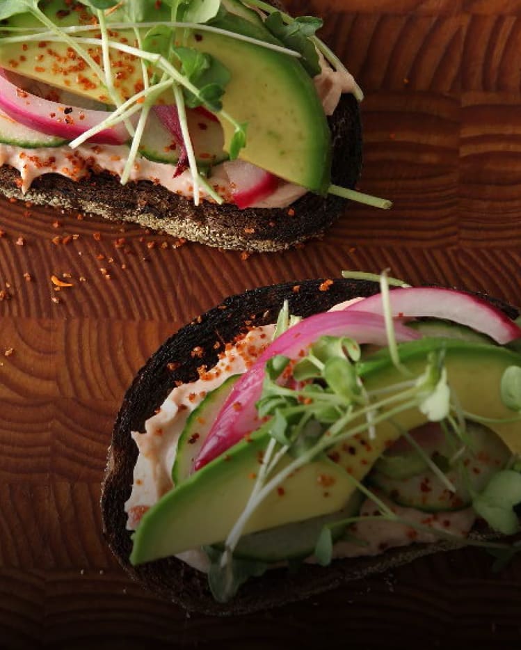 Two slices of dark rye toast topped with Philadelphia cream cheese, sliced avocado, cucumbers, pickled onions, microgreens.