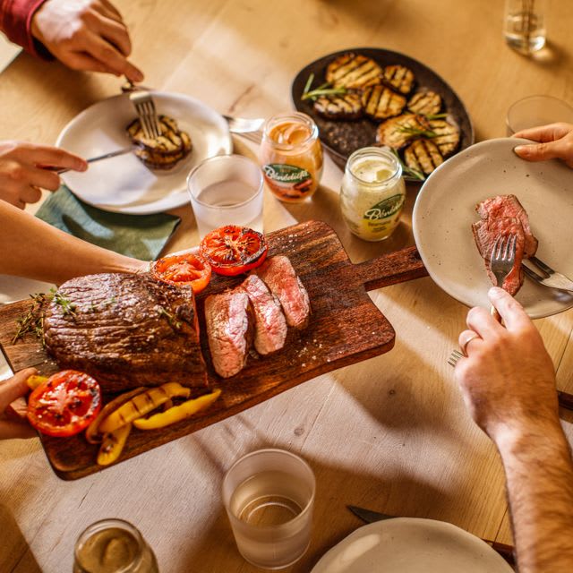 Image d'une table garnie de nourriture, notamment du steak, des tomates et des personnes mangeant.