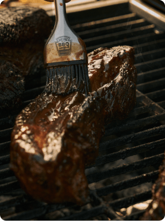 ハインツのシリコンブラシを使い、網の上で焼くジューシーな肉の塊にBBQソースを塗る様子
