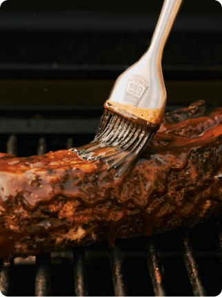 ハインツのシリコンブラシを使い、網の上で焼くジューシーな豚バラ肉にBBQソースを塗る様子。