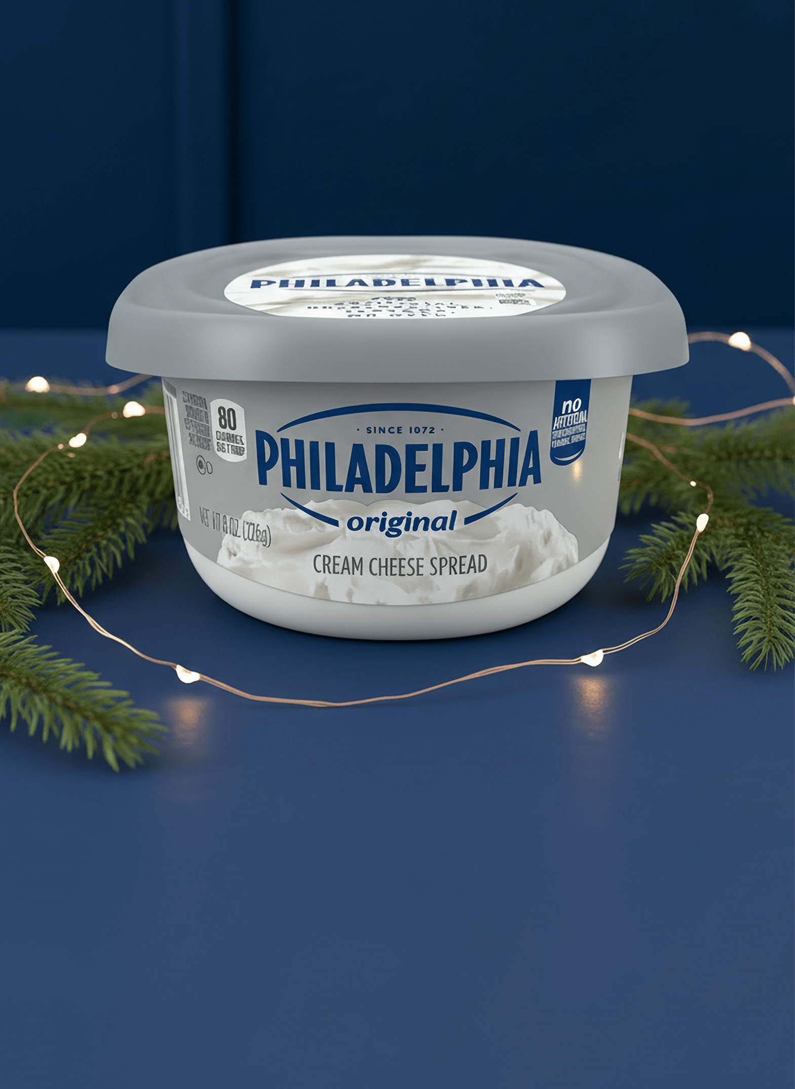  A tub of Philadelphia Original Cream Cheese Spread on a dark blue surface, surrounded by pine boughs and string lights.