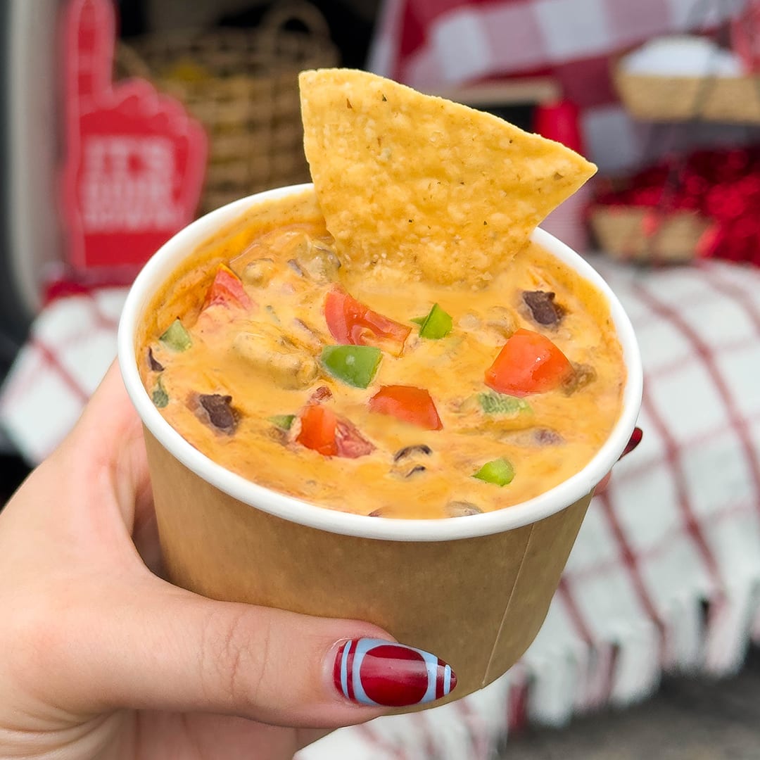 A hand holding a cup of creamy Velveeta Cowboy Queso dip, loaded with vibrant red tomatoes, green bell peppers
