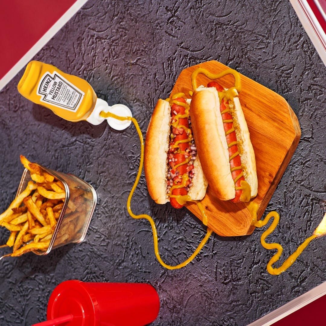 Two hot dogs topped with Heinz Yellow Mustard and diced onions on a wooden serving board with french fries on the side and a 