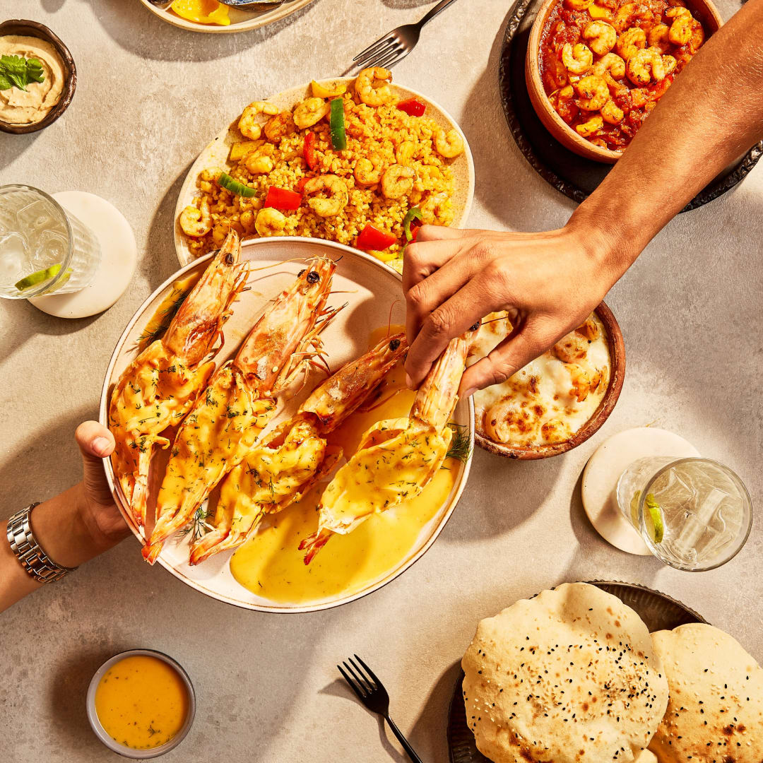 Overhead view of a table with various seafood dishes, including grilled shrimp, bulgur, bread, hummus, and drinks.