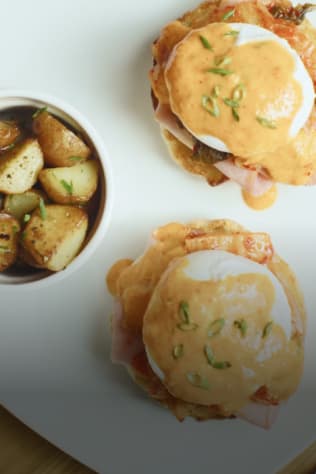 Two Eggs Benedict with a side of roasted potatoes, drizzled with Hollandaise sauce and garnished with fresh herbs.