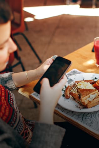 A woman looking at her phone while sitting at a table with a plate of sausage, eggs, and toast. A glass of red juice is nearb