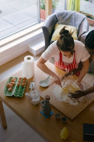 Overhead shot of two people preparing dough on a wooden cutting board in a bright kitchen.