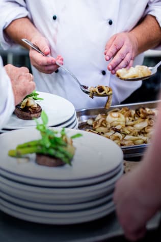Chefs plating a dish with steak, asparagus and sauteed onions in a busy kitchen.