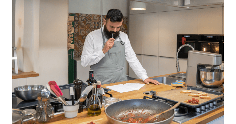 Bearded man in apron ponders recipe in kitchen.