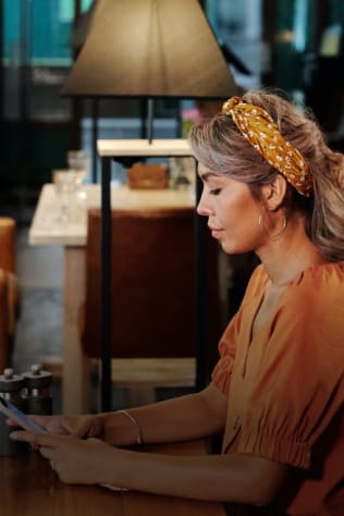 A woman sitting at a restaurant table looking at her phone. She is wearing an orange shirt and a patterned headband.