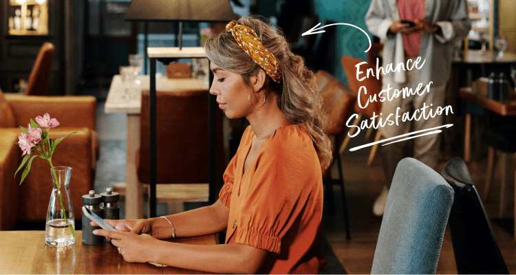 Woman sitting down in a restaurant using her cellphone with a man in the background standing by the bar using his cellphone