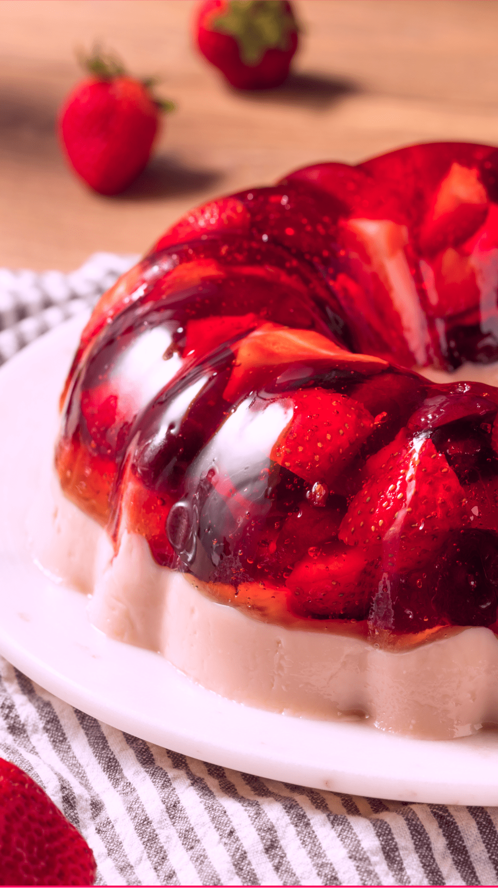 A glistening, red Jell-O mold, studded with strawberries and blueberries, sits on a pristine white plate. The mold rests on a