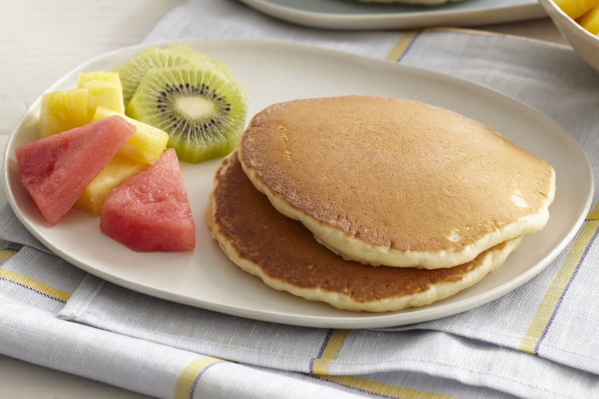 Two fluffy golden-brown pancakes served with sliced kiwi, pineapple, and watermelon.
