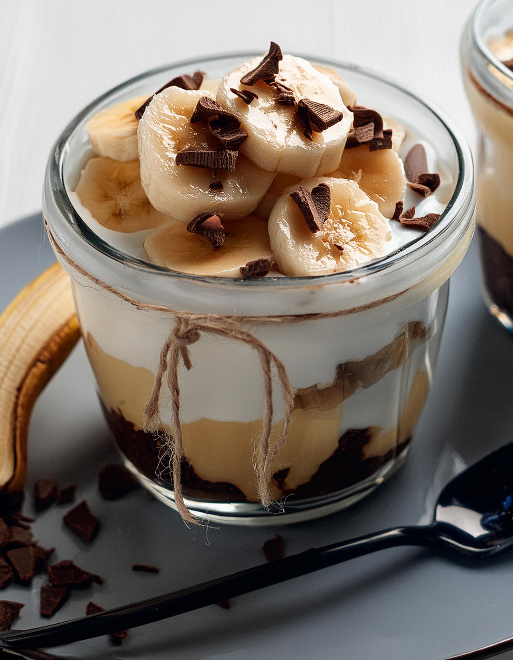 Toetje in een glas met laagjes banaan, chocolade en room, afgewerkt met chocoladeschilfers.