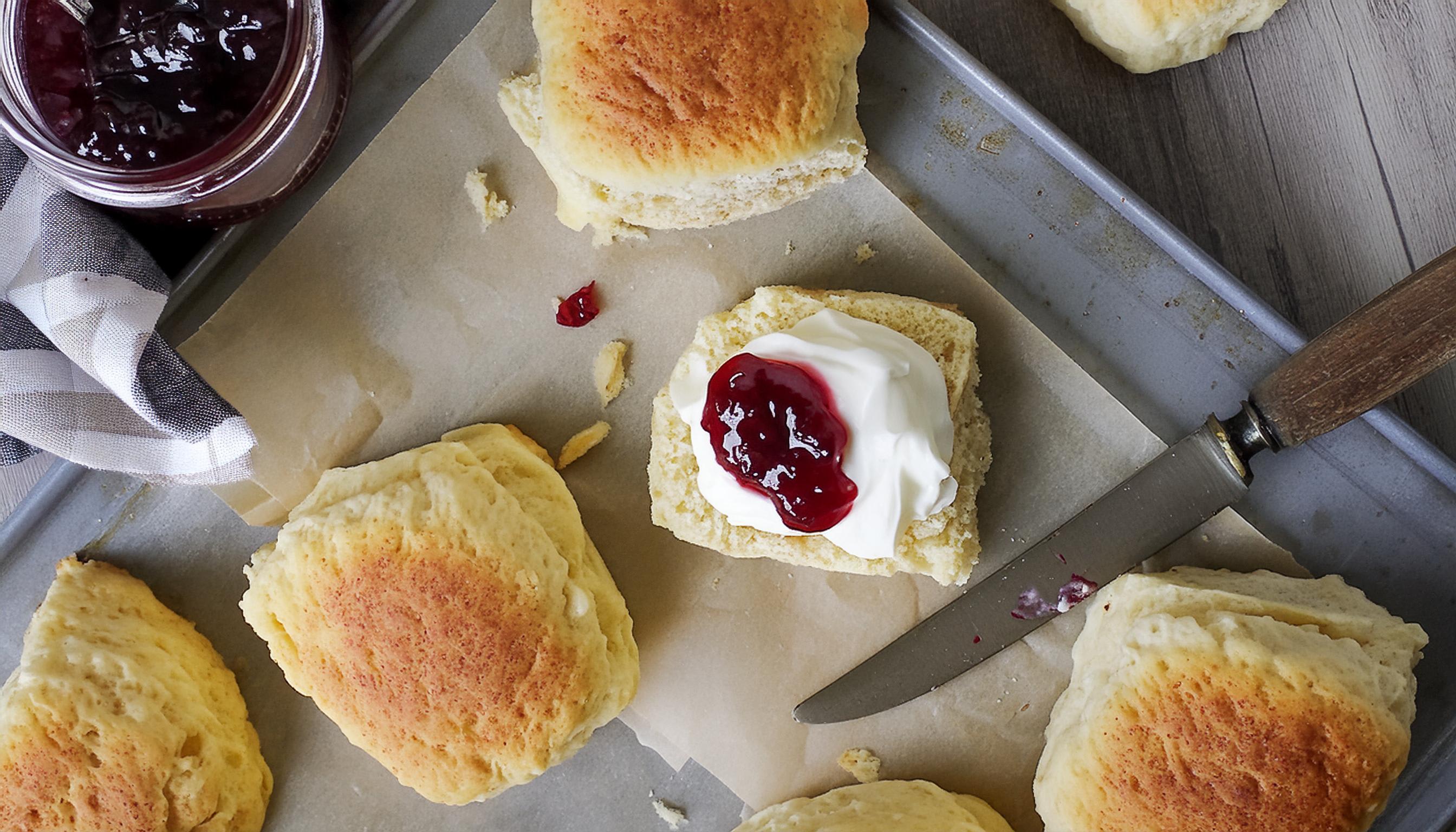 Fluffy scones on a tray, one topped with whipped cream and red jam.