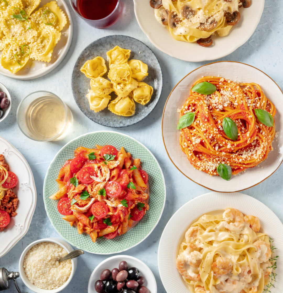 A tabletop spread of different pastas and sauces, with parmesan cheese and olives.
