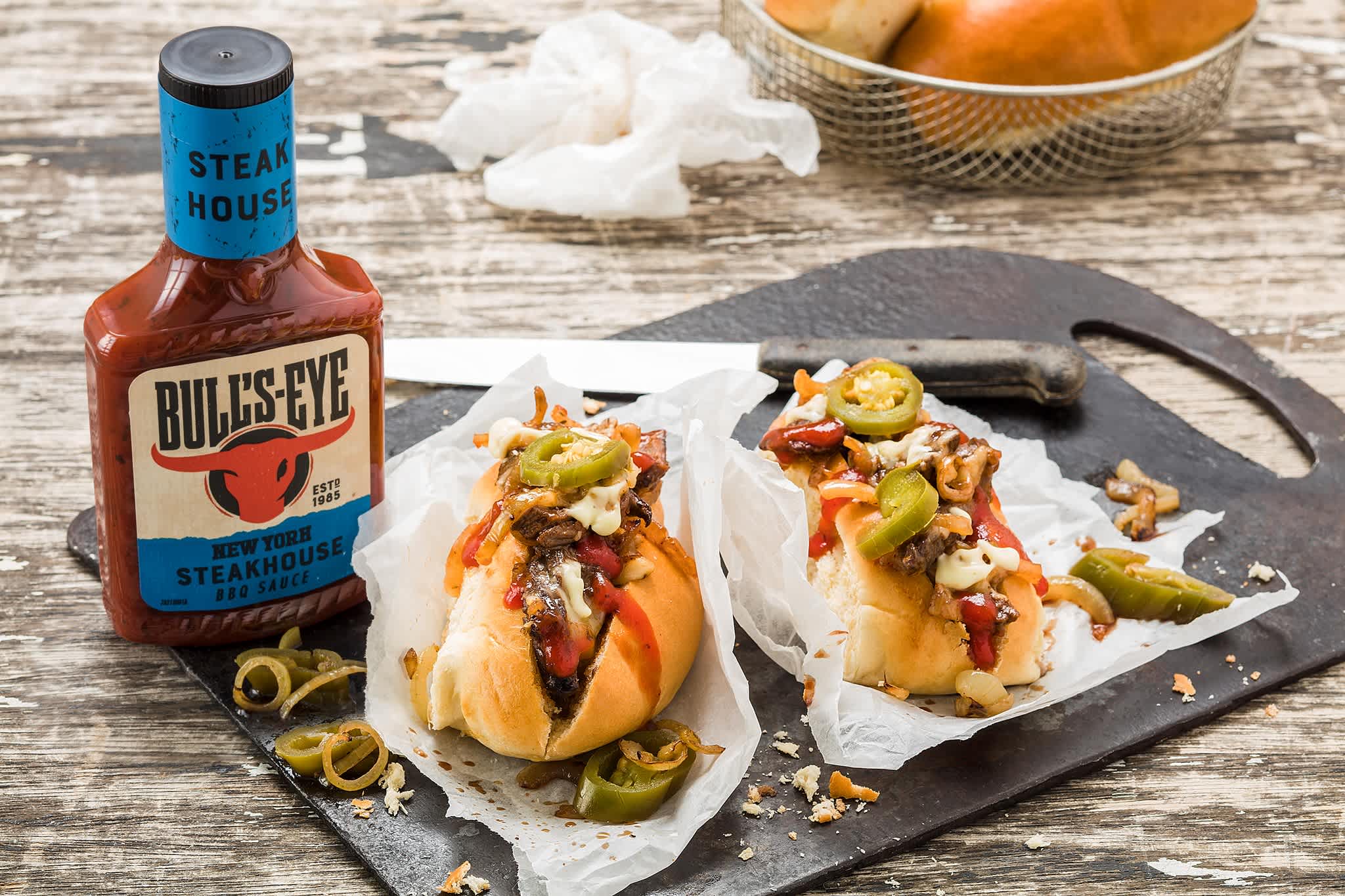 Loaded steak subs with melted cheese and jalapeños on a black board with BBQ sauce.