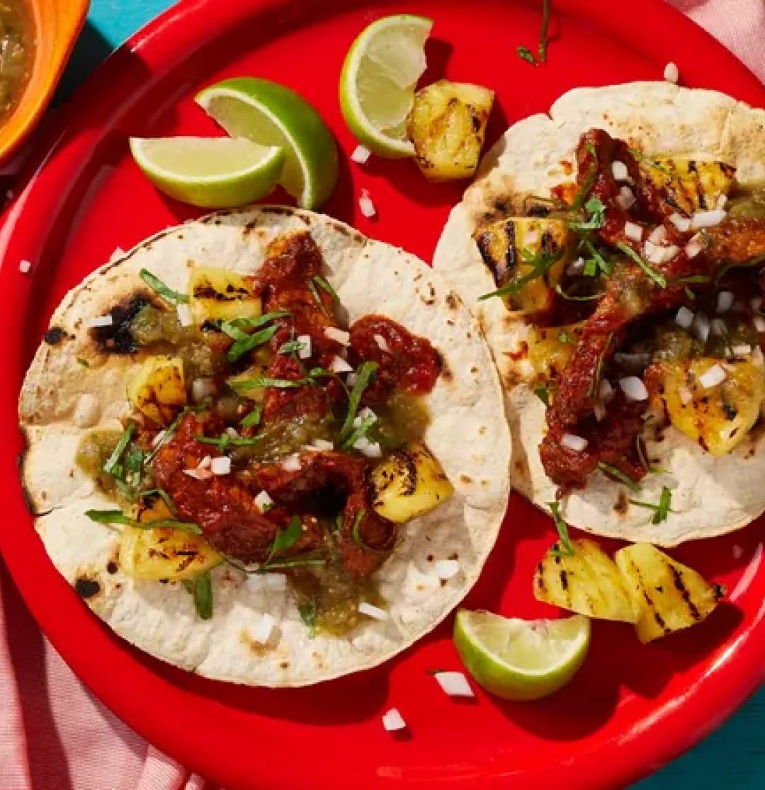 Two tacos with pineapple, meat, salsa, and lime on a red plate.