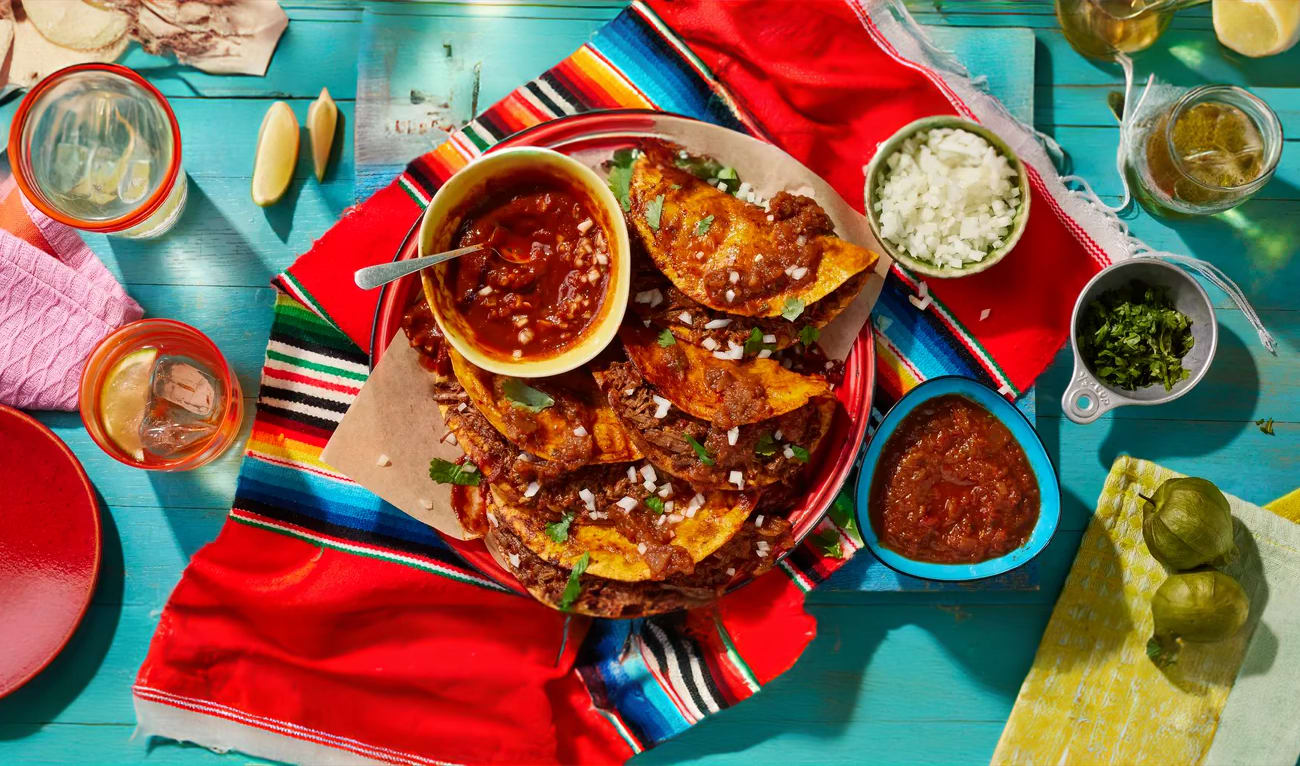 A platter of folded, crispy beef tacos served with a bowl of consommé for dipping.