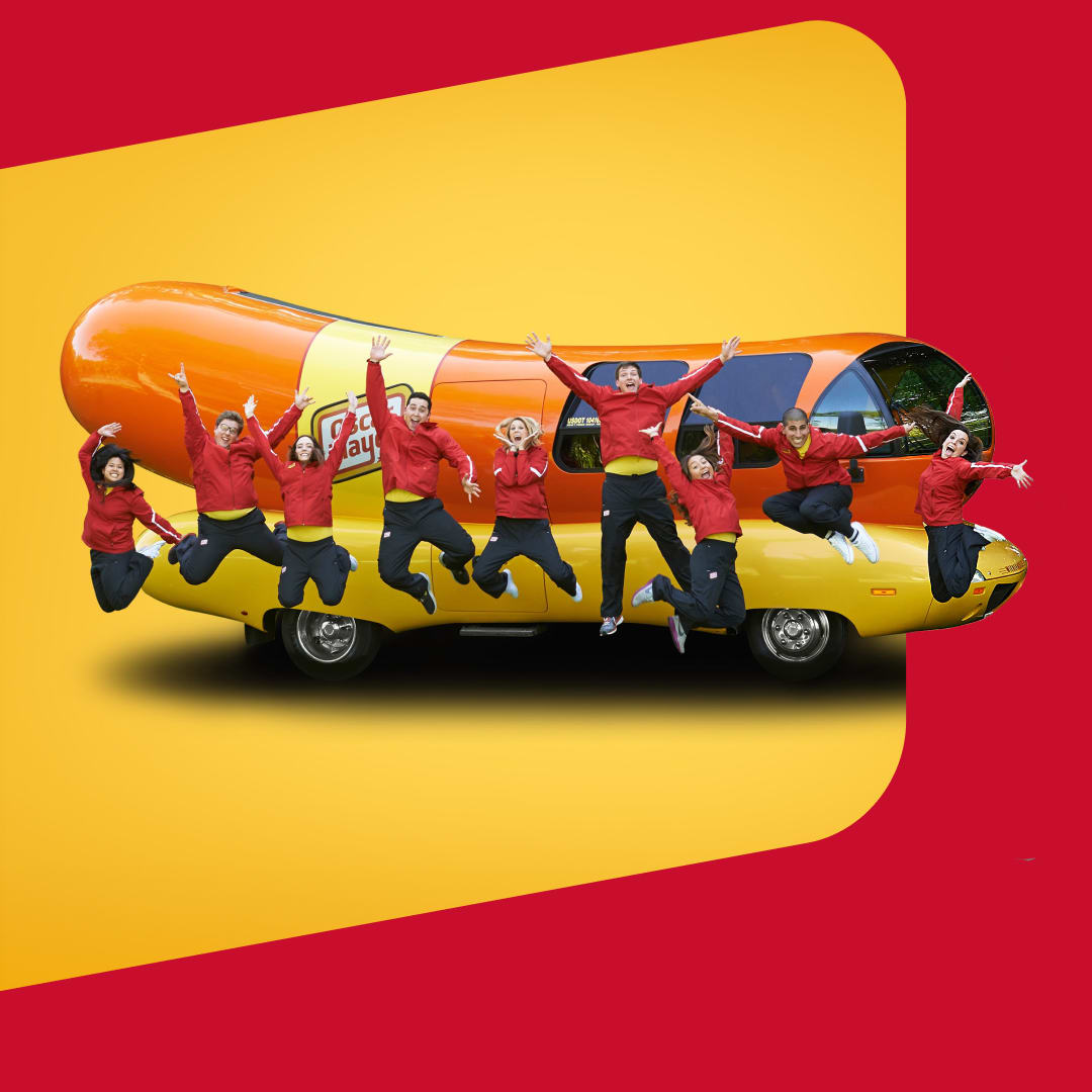 The energetic Oscar Mayer team jumps for joy in front of the Wienermobile, a giant hot dog-shaped vehicle.