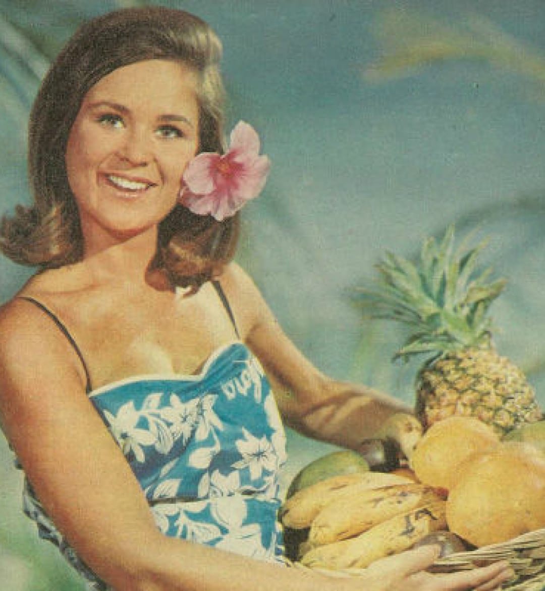 An old photo of a woman wearing a floral dress carrying a basket of fruit.
