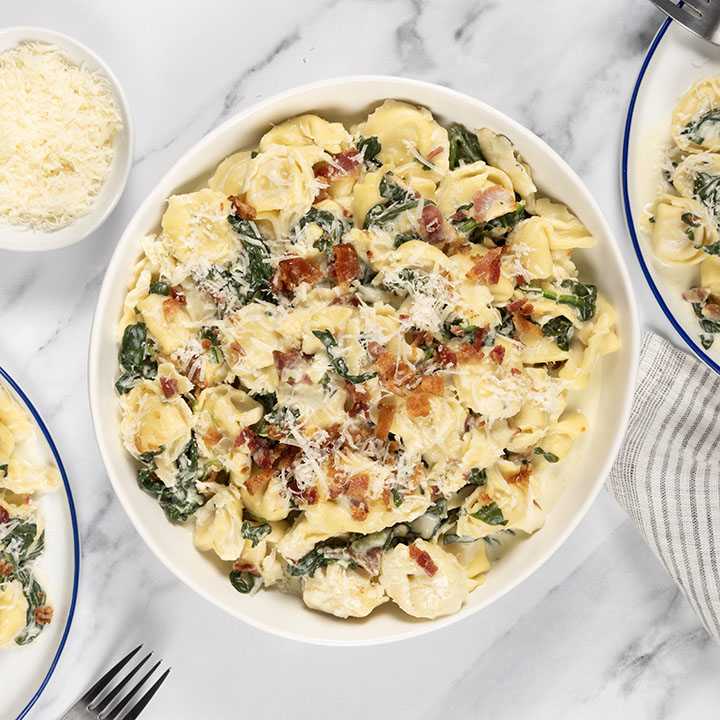 A top-down view of a bowl of tortellini alfredo, generously topped with crispy bacon bits and fresh spinach. Grated parmesan cheese adds a final touch to this decadent dish. Two smaller plates with servings of the same dish, a bowl of parmesan cheese, and silverware are also visible on the marble surface. A touch of texture is present with a striped napkin in the background.