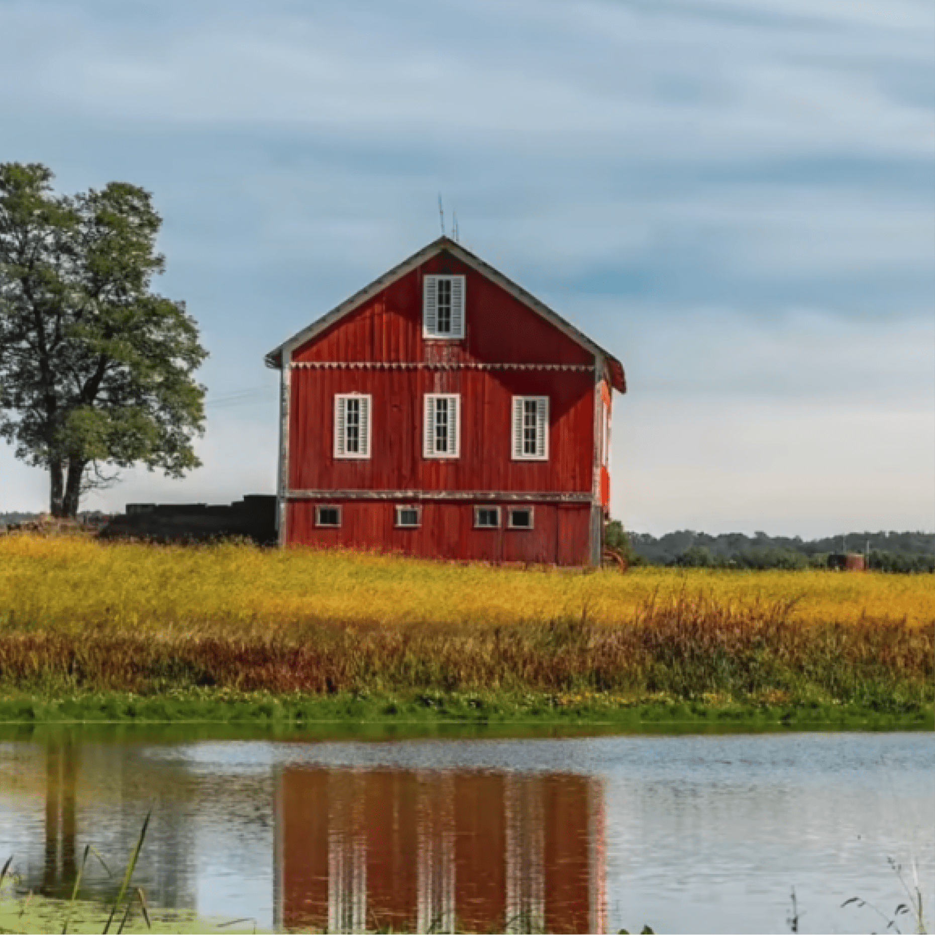 A bright red barn with white-trimmed windows stands in a golden field beside a calm body of water. Its reflection is visible 