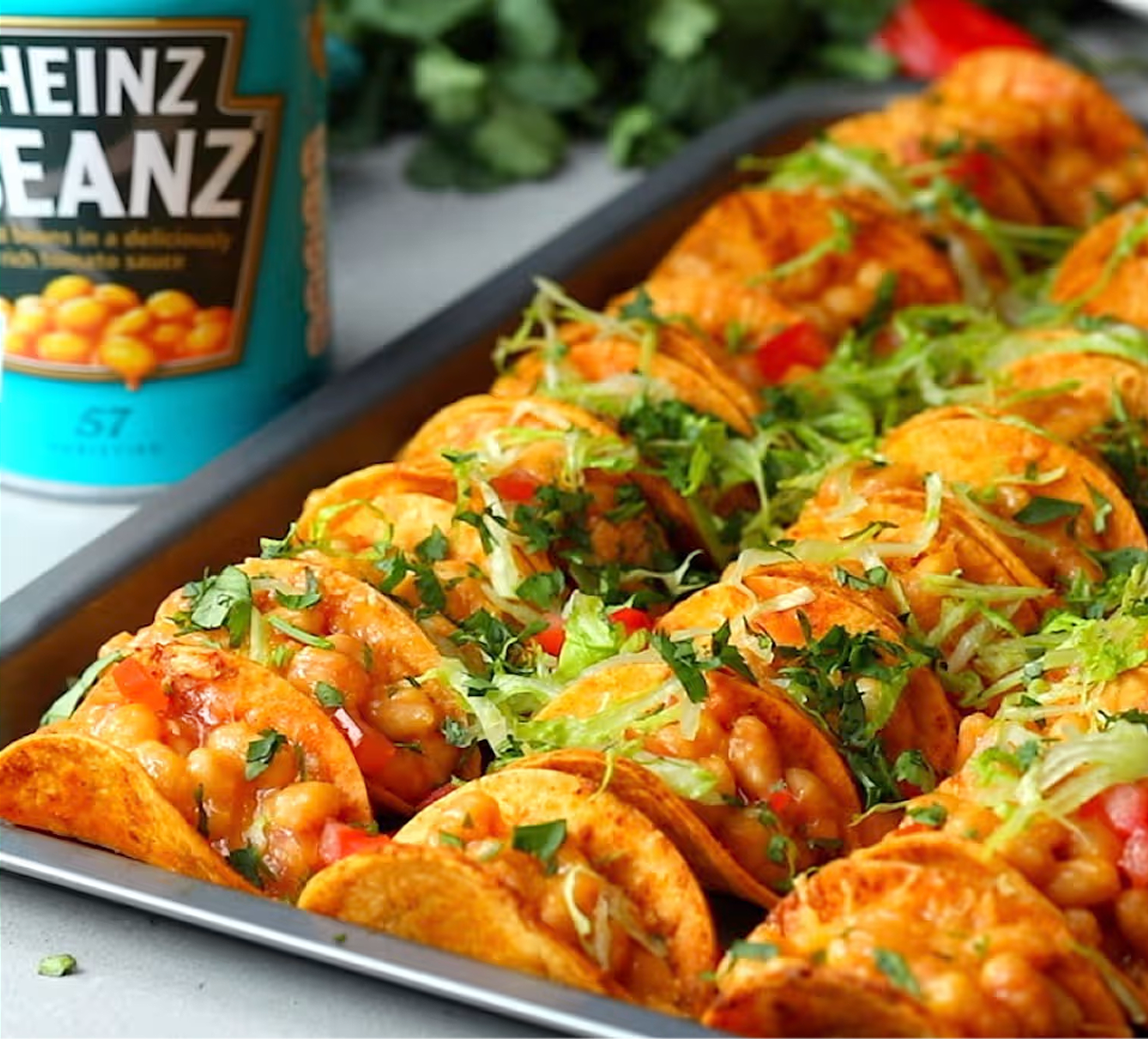 Na blasze do pieczenia ułożone są mini tacos z fasolą Heinz, sałatą, pomidorami, serem i ziołami. W tle widoczna jest puszka 