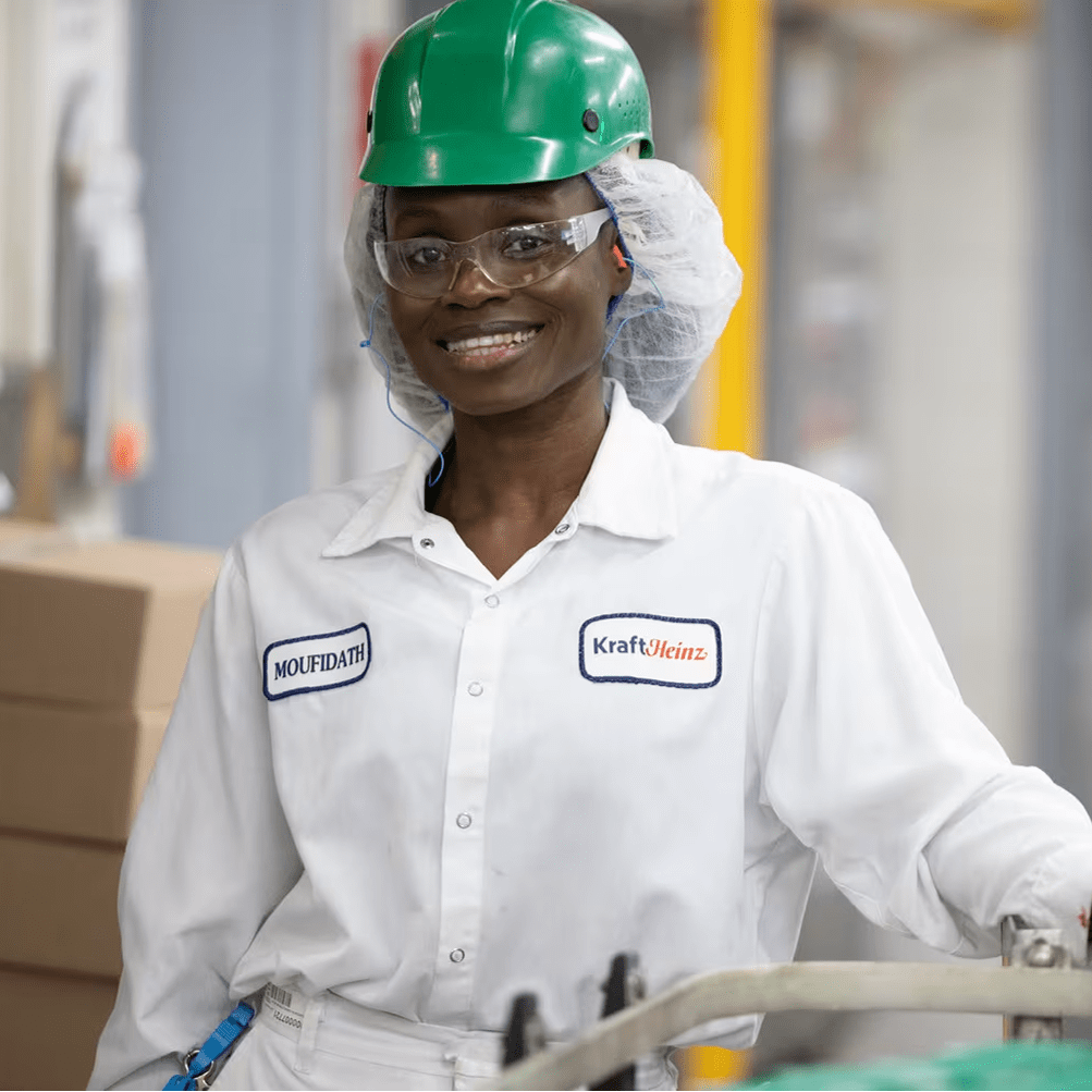 Femme en usine Kraft Heinz, casque vert, lunettes et uniforme, souriante et s'appuyant sur un équipement industriel.