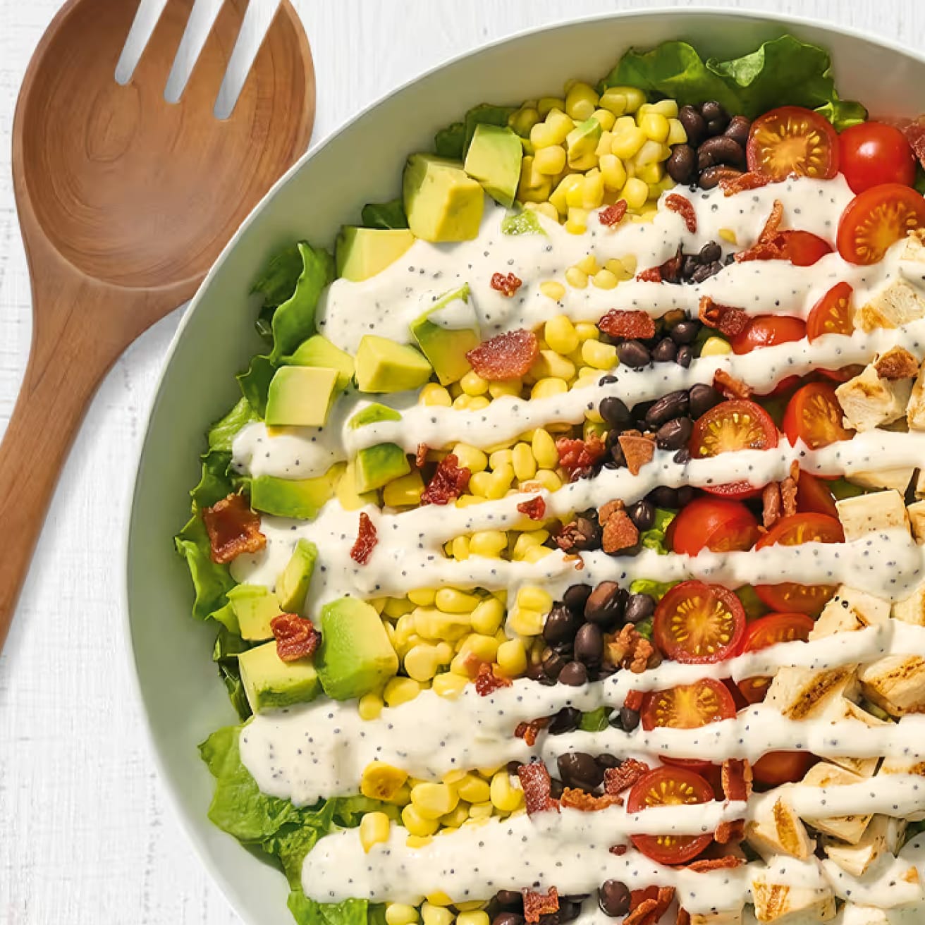 Garden salad bowl with rows of avocado, corn, beans, tomatoes, chicken, and bacon with creamy dressing.