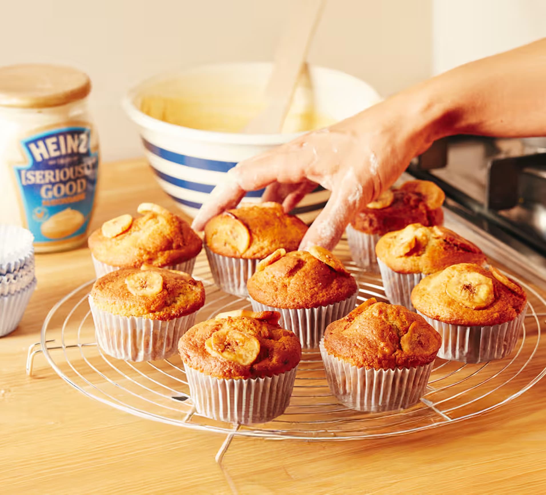 Golden-brown banana muffins cooling on a wire rack, made with a secret ingredient: Heinz "Seriously Good" Mayonnaise. A jar o