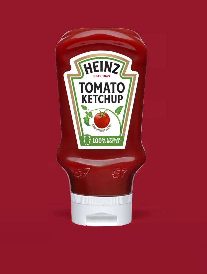 Una botella de ketchup de tomate Heinz con tapón blanco, sobre un fondo rojo liso. En la etiqueta se lee "HEINZ ESTD 1869 TOM