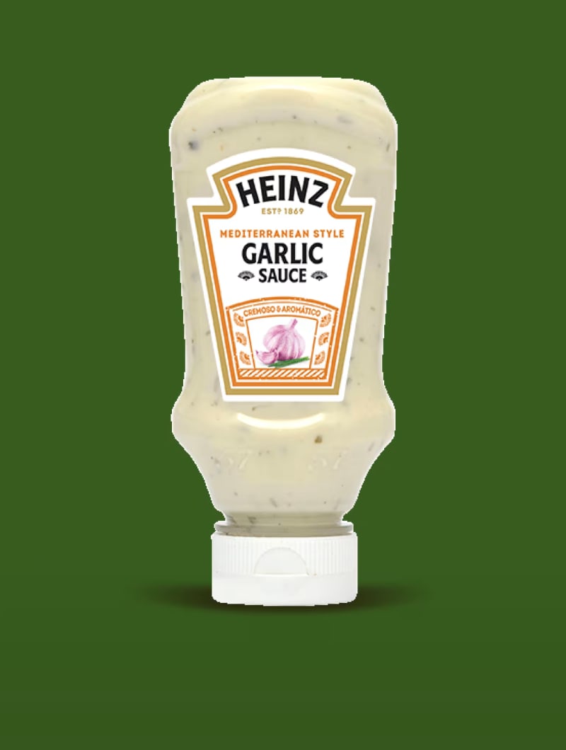 Düz, orta yeşil bir arka planın ortasında baş aşağı duran bir Heinz Akdeniz Stili Sarımsaklı Sos sıkma şişesi.