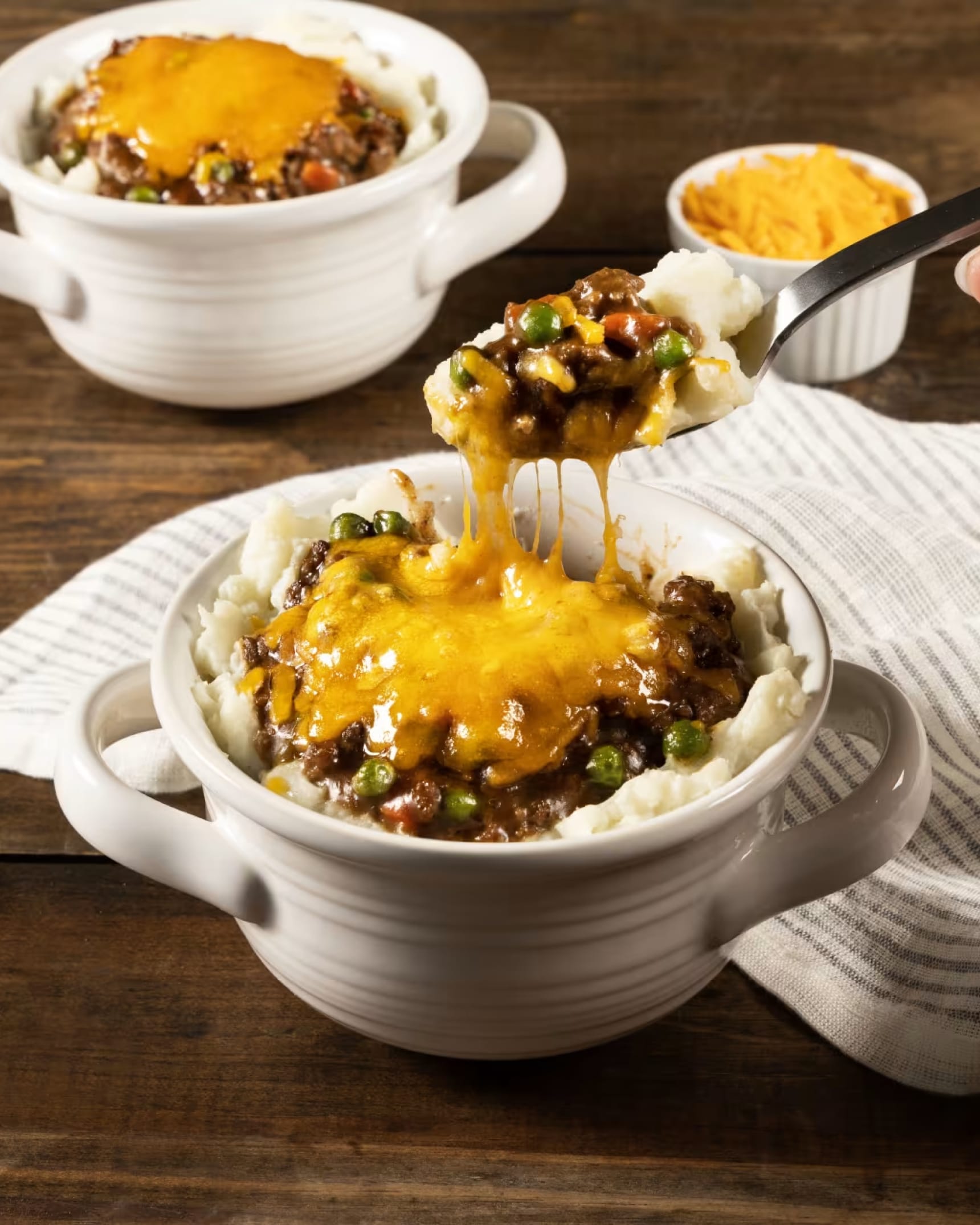 A spoonful of shepherd's pie with melted cheddar cheese pulling from a ceramic bowl on a wooden table.