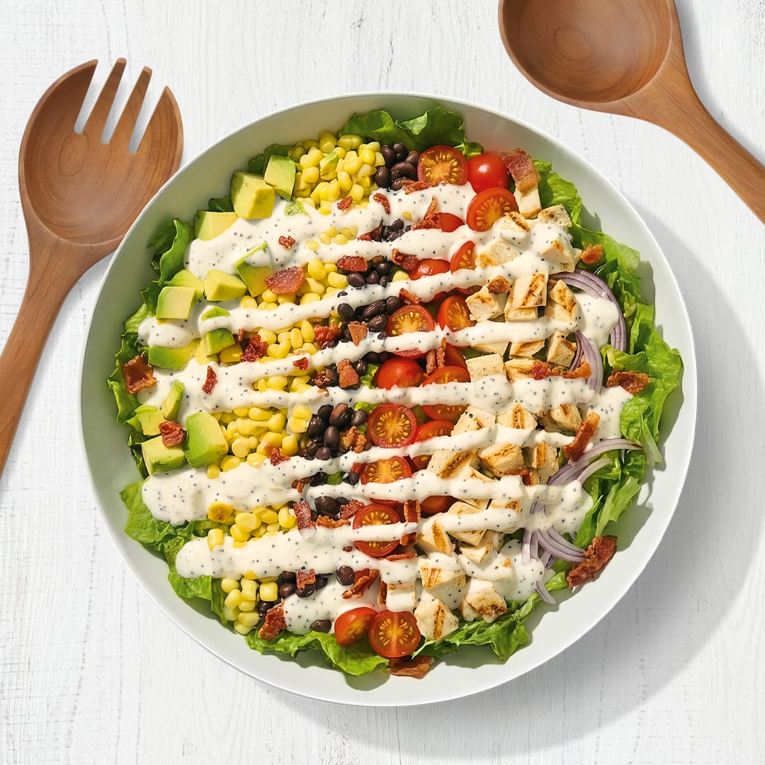 A vibrant chicken salad bowl with rows of fresh vegetables and creamy Kraft Classic Ranch dressing on a white surface.