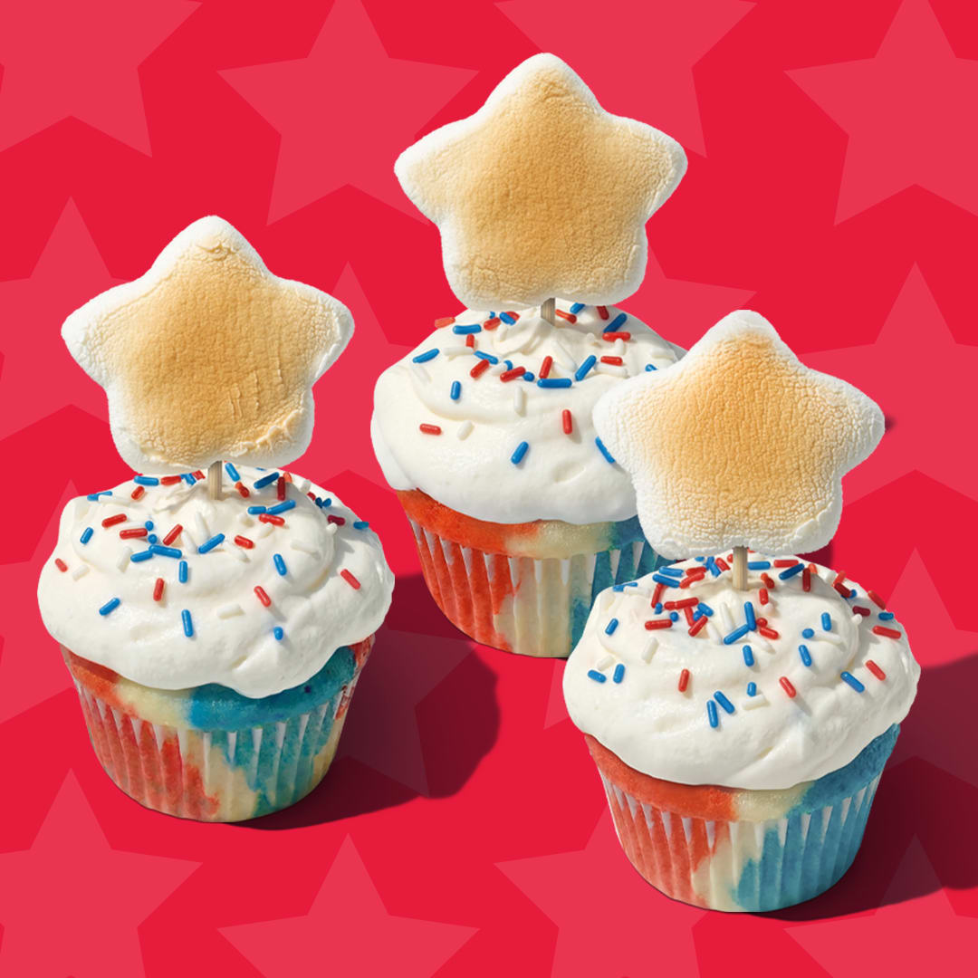 Red, white, and blue swirled cupcakes topped with sprinkles and star marshmallows against a red star background.