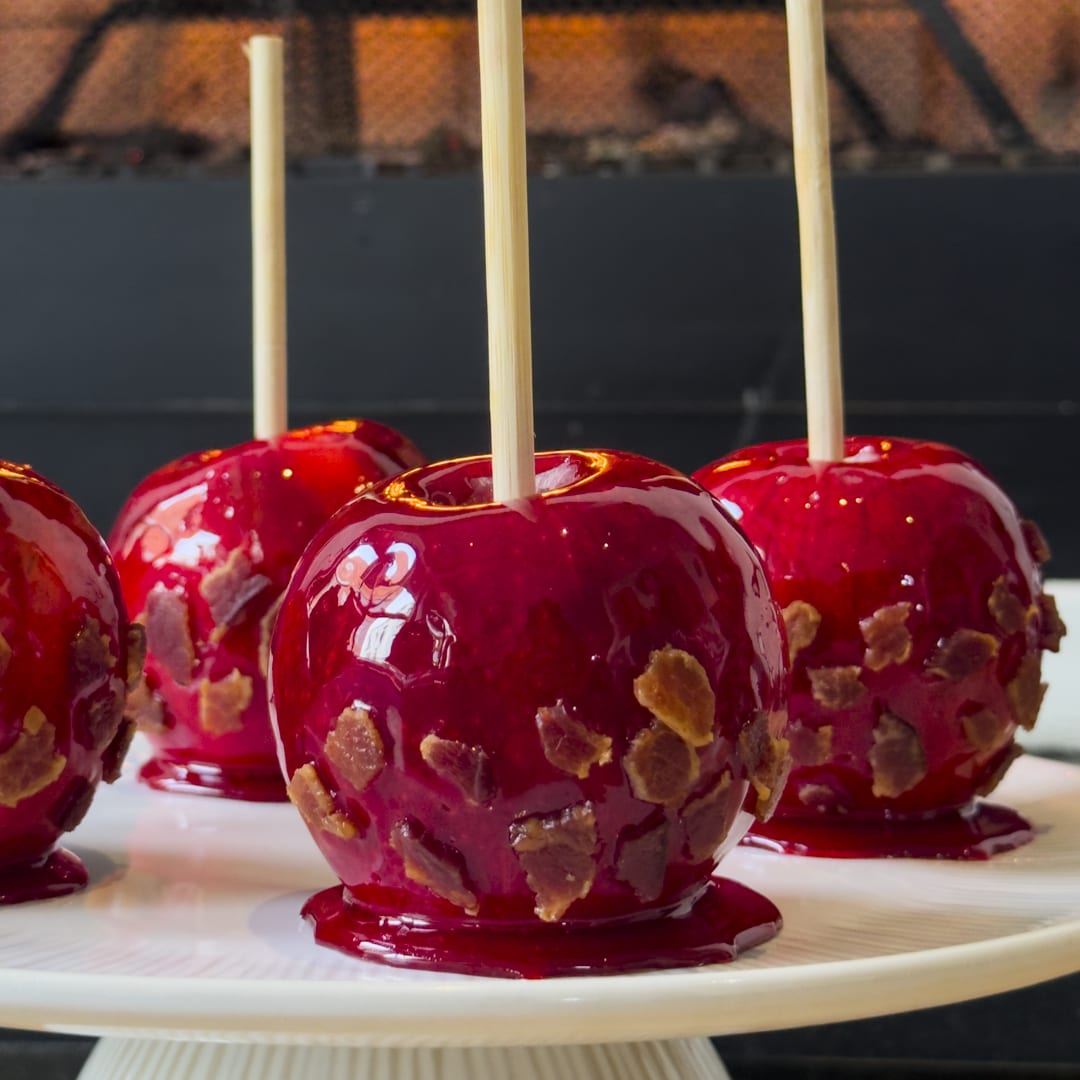 Deep red candied apples on wooden sticks, coated with golden-brown bacon bits, arranged on a white fluted cake stand. 