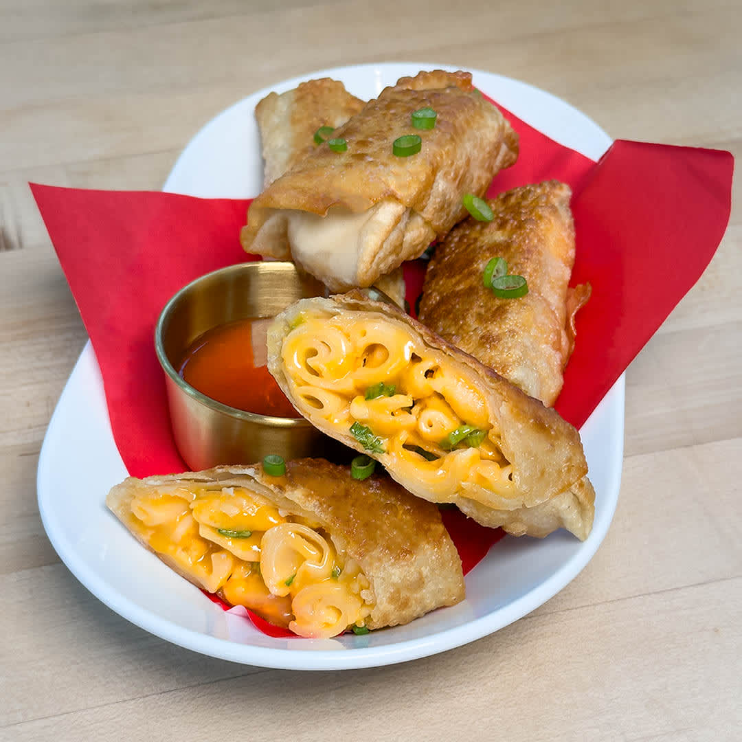 Four crispy Velveeta Cheesy Mac & Cheese Egg Rolls are displayed on a white plate with a red napkin. Two of the egg rolls have been cut in half to show the cheesy mac & cheese filling. A small gold bowl of dipping sauce sits next to the egg rolls.