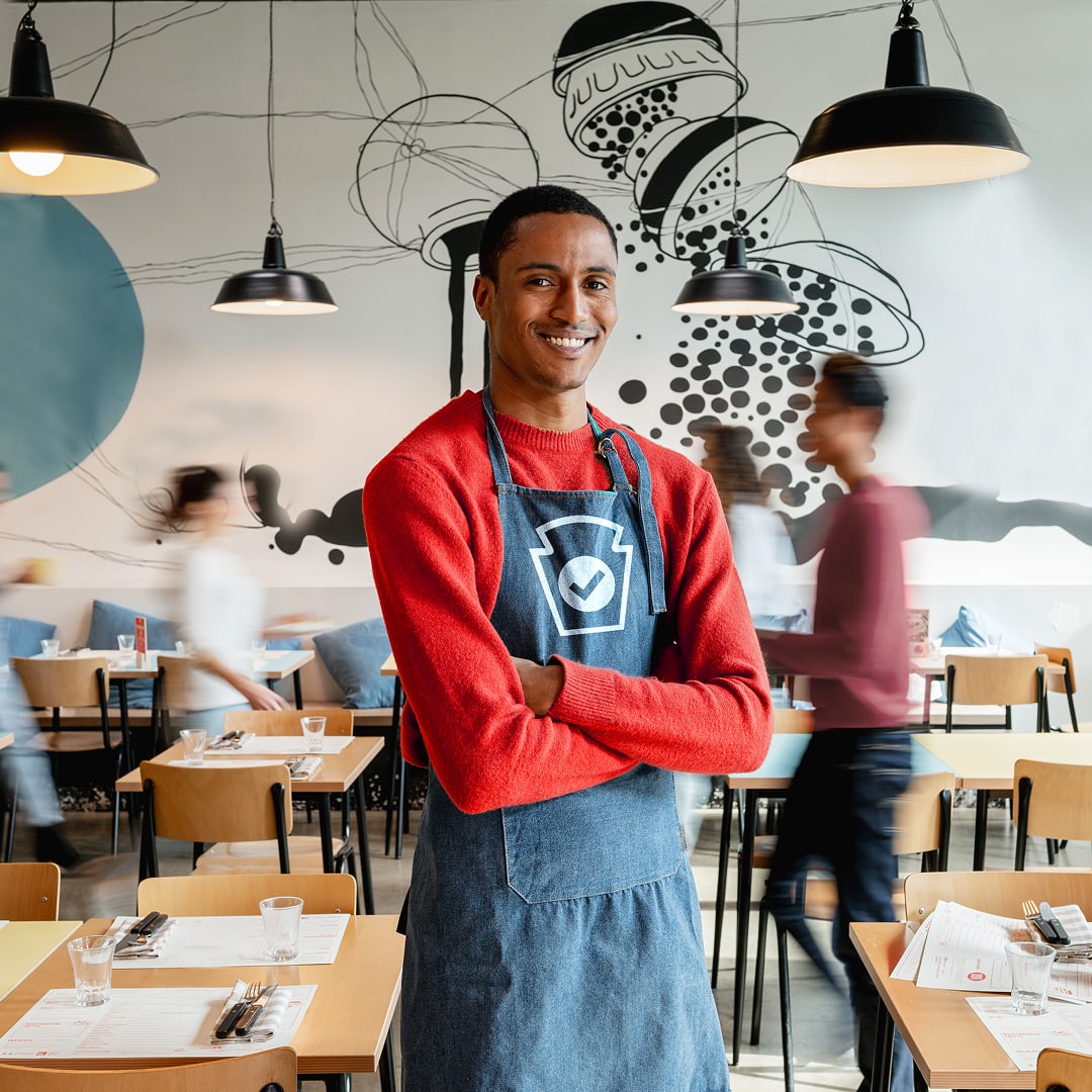 A smiling server wearing a Heinz Verified apron stands in a modern, bustling restaurant.
