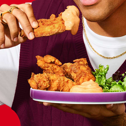 Close-up of a person wearing a maroon sweater vest and gold chain, about to enjoy a crispy chicken tender dipped in a creamy, pinkish sauce. They hold a plate filled with more chicken tenders, a generous portion of the same sauce, and a side salad. Against a vibrant red background, this image represents the "Appetizer" filter for recipes.