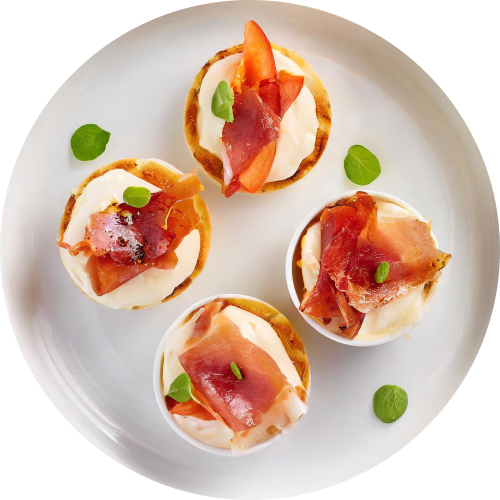 Overhead shot of four miniature potato pancakes topped with cream cheese, thinly sliced prosciutto, a small piece of tomato, and a garnish of fresh oregano.
