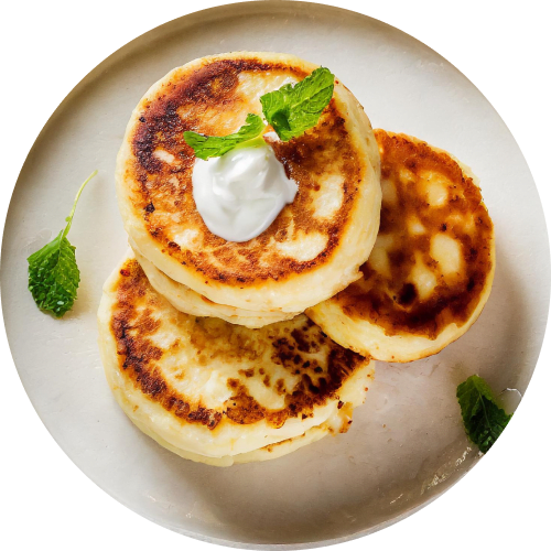 Overhead close-up of three golden-brown cheese pancakes (syrniki) stacked on a plate. They're topped with a dollop of sour cream and garnished with fresh mint leaves.