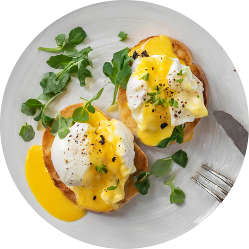 Overhead close-up of two eggs Benedict on a white plate. Each English muffin half is topped with a poached egg, creamy hollandaise sauce, fresh watercress, and a sprinkle of black pepper. A fork and knife are placed beside the dish.
