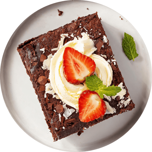 Overhead shot of a fudgy brownie square on a plate, adorned with a dollop of whipped cream, two fresh strawberry halves, a sprig of mint, and chocolate shavings.