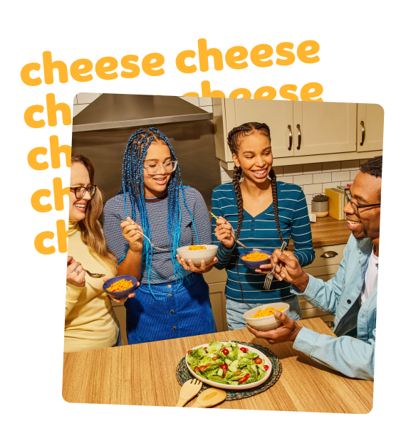 A family laughs around a kitchen island for a weeknight dinner, eating bowls of Kraft Mac and Cheese with a tossed salad