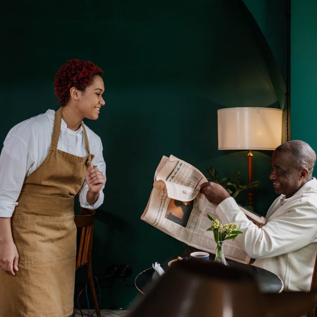 Chef in a brown apron standing up talking to a customer sitting down