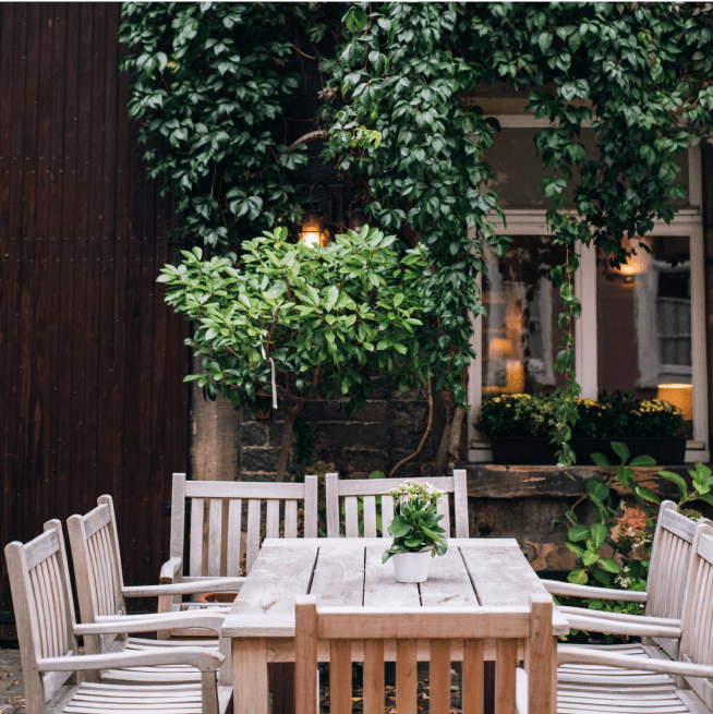 Wooden tables and chairs outside in front of a brick wall with plants